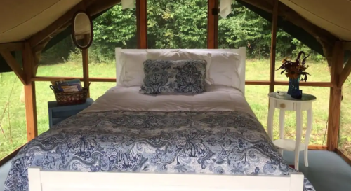 Cozy bedroom in a tent with a white bed, blue patterned bedding, and a view of greenery outside.