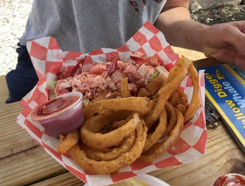 A basket of lobster roll with onion rings and a side of dipping sauce on a wooden table.