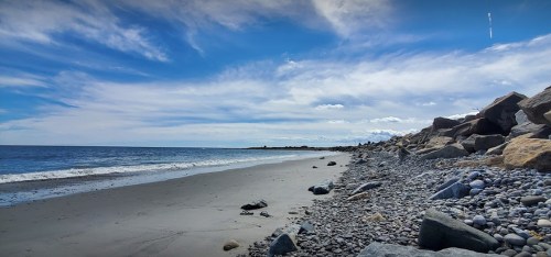 A serene beach scene with smooth sand, pebbles, and gentle waves under a blue sky with scattered clouds.