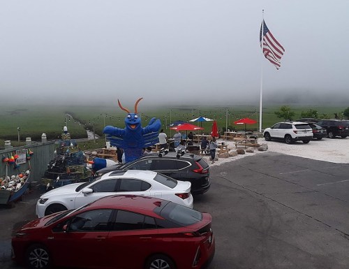 A foggy scene at a seafood restaurant with a large blue lobster statue, cars parked, and people dining under umbrellas.