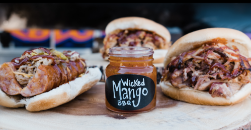 Two BBQ sandwiches with pulled meat, topped with coleslaw, beside a jar of Wicked Mango BBQ sauce on a wooden board.