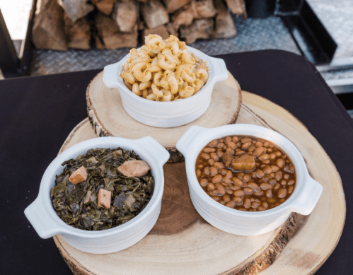 Three bowls of food on a wooden platter: macaroni and cheese, collard greens with sausage, and baked beans.