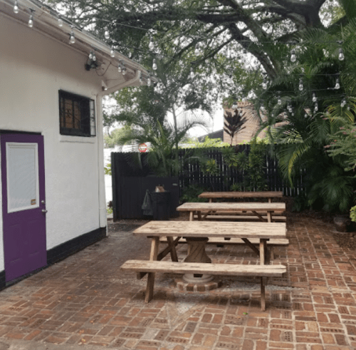 Outdoor seating area with wooden picnic tables, surrounded by greenery and string lights, next to a white building.