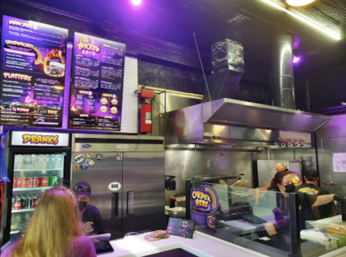 A vibrant food counter with a menu board, drinks fridge, and staff preparing food in a modern eatery.