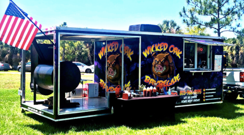 A food truck named "Wicked Oak Barbecue" with a large smoker, condiments, and an American flag in a grassy area.