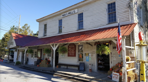 Historic building with a red roof, sign reading "Nora Mills," and autumn decorations outside. American flag displayed.