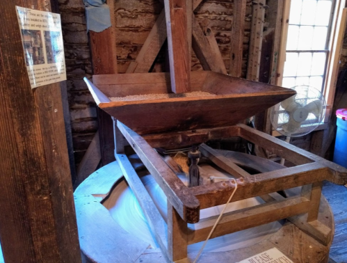 A wooden grain mill with a hopper, set in a rustic interior with exposed beams and a fan in the background.