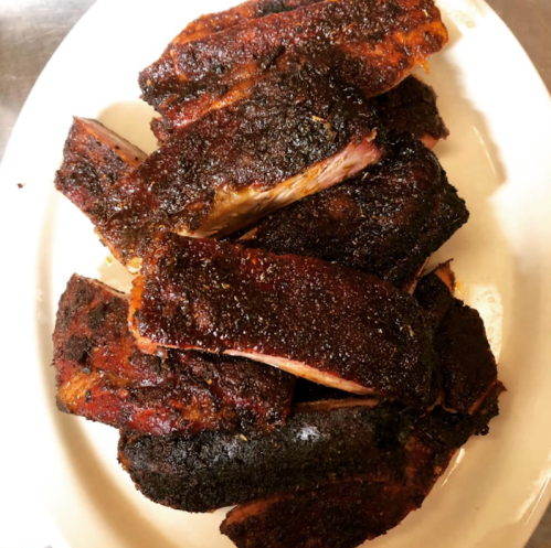 A platter of smoky, charred ribs with a dark, flavorful crust, arranged neatly on a white plate.