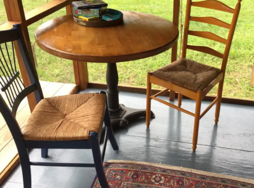 A wooden table with two chairs, one blue and one natural, on a porch with a grassy view outside.