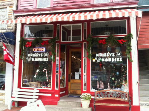 A festive candy shop with a red exterior, decorated for the holidays, featuring an "OPEN" sign and window displays.