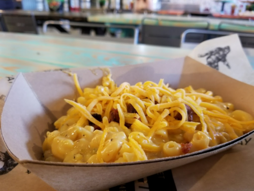 A serving of creamy macaroni and cheese topped with shredded cheddar in a paper tray on a colorful table.