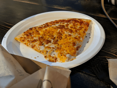 A slice of cheese pizza on a white paper plate, with melted cheese and a golden crust, resting on a wooden table.