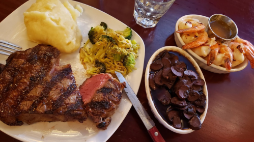 A plate with grilled steak, mashed potatoes, broccoli, noodles, and a side of shrimp and mushrooms.