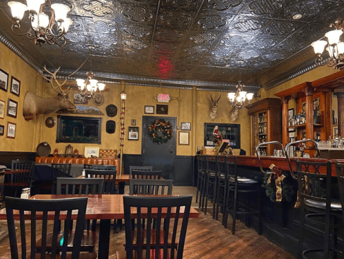 Cozy restaurant interior with wooden tables, a bar, festive decorations, and vintage decor including a mounted deer head.