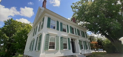 A white historic house with green shutters, surrounded by trees and a blue sky with fluffy clouds.