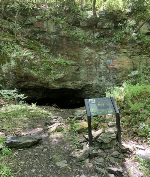 A rocky cave entrance surrounded by greenery, with a sign labeled "Marble Hill" in the foreground.