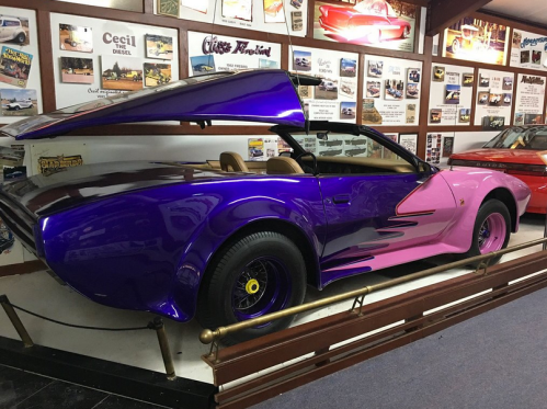 A vibrant purple and pink custom car displayed in a museum, featuring a unique design and open rear wing.