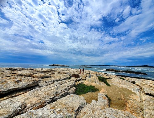 Two people walking on rocky shoreline under a blue sky with scattered clouds and ocean waves in the background.