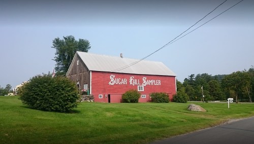 Red barn with "Sugar Hill Sampler" painted on the side, surrounded by green grass and trees. Clear blue sky above.
