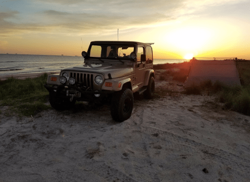 A Jeep parked on a sandy beach at sunset, with a tent nearby and the ocean in the background.