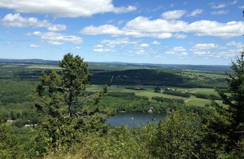 A scenic view from a hilltop, showcasing lush green fields, a winding river, and a blue sky with fluffy clouds.