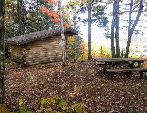 A wooden cabin beside a picnic table, surrounded by colorful autumn trees and a scenic view in the background.