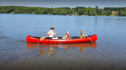 A man and two children paddle a red canoe on a calm lake surrounded by green trees.