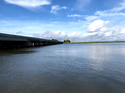 A calm river scene with a bridge overhead and blue skies dotted with clouds in the background.