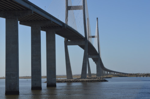 A large suspension bridge spans over a calm river, with tall pillars and cables supporting its structure against a clear sky.