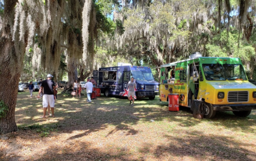 Food trucks parked under trees in a park, with people walking and enjoying the outdoor atmosphere.