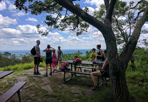 A group of hikers resting at a scenic overlook, surrounded by trees and a view of the mountains under a cloudy sky.