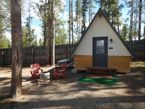 A small A-frame cabin surrounded by trees, with red chairs and a picnic table in a gravel area.
