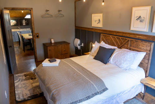 Cozy bedroom with a wooden bed, gray walls, and warm lighting, featuring a nightstand and a view into a dining area.