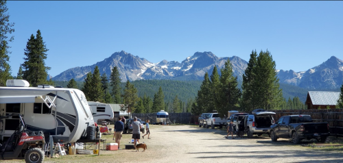 A scenic campsite with RVs, people, and a dog, set against a backdrop of snow-capped mountains and clear blue skies.