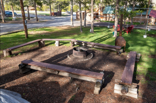A circular fire pit surrounded by wooden benches in a grassy area, with trees and cabins in the background.