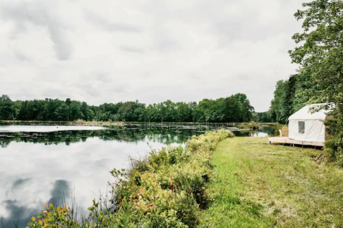 A serene lakeside scene with a glamping tent, surrounded by lush greenery and a cloudy sky reflecting on the water.
