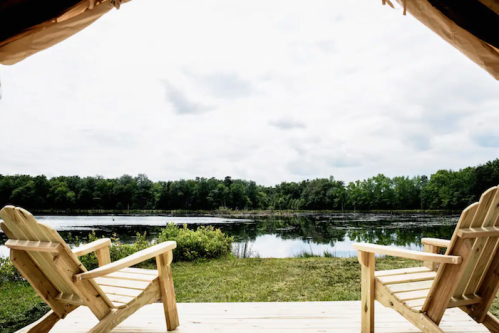 Two wooden chairs face a serene lake surrounded by greenery under a cloudy sky.