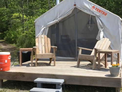 A cozy glamping tent with wooden chairs on a deck, surrounded by greenery. A cooler and potted flowers are nearby.