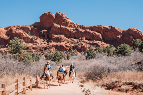 Three riders on horseback traverse a dirt path with red rock formations and trees in the background under a clear blue sky.