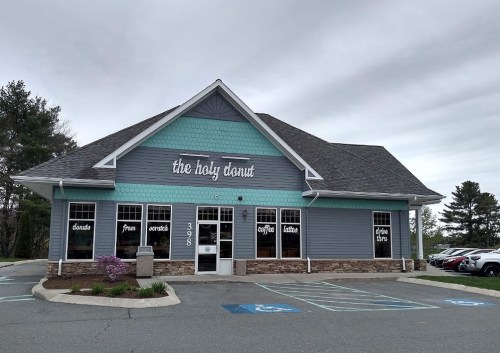 Exterior of "The Holy Donut" shop, featuring a blue and gray facade, large windows, and a parking lot.