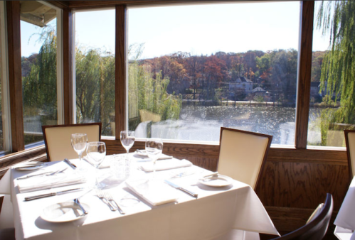 A dining table set for four, with a scenic view of a lake and autumn trees through large windows.