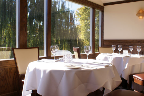 A cozy restaurant interior with a round table set for dining, featuring glassware and large windows with a view.
