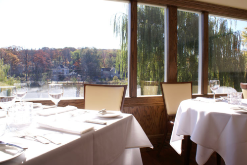 Elegant dining setup with white tablecloths and glassware, overlooking a scenic view of trees and water.