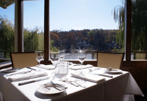 A beautifully set dining table with glassware, overlooking a scenic view of trees and a lake through large windows.