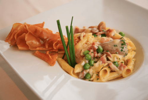 A plate of creamy pasta with chicken, peas, and bacon, accompanied by crispy potato chips and garnished with chives.