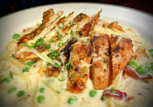 Creamy pasta with grilled chicken, peas, and crispy bacon, garnished with herbs and cheese.