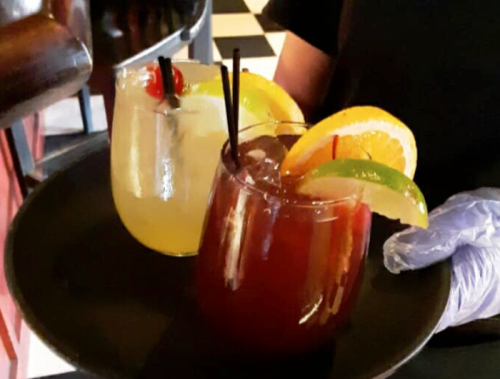 A server holds a black tray with two colorful cocktails garnished with fruit slices and cherries.