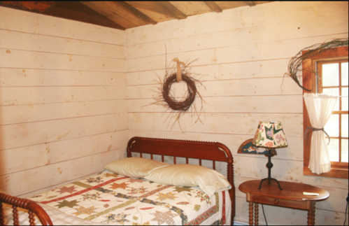 Cozy bedroom with a wooden bed, quilted bedding, a small table, and a rustic wreath on the wall. Natural light from a window.