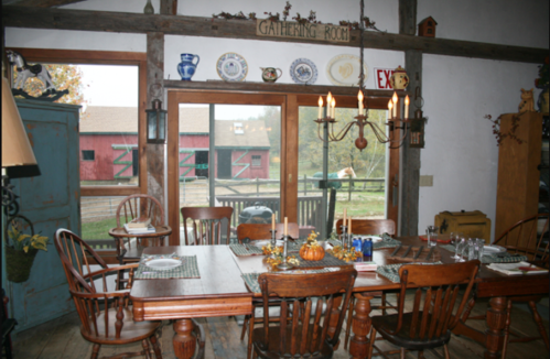 Cozy dining room with a wooden table, chairs, and a view of a barn through large windows. Decorated for a gathering.