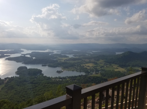 A scenic view of a lake surrounded by hills and greenery, with clouds in a blue sky. A wooden railing in the foreground.